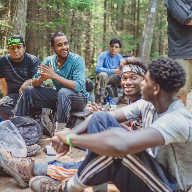 The image shows a group of young men sitting in a wooded area, possibly a park or forest. They appear to be engaged in conversation and seem relaxed. Some are sitting on the ground, while others are on what looks like makeshift seats. The atmosphere seems casual and friendly, suggesting they are enjoying each other's company in an outdoor setting.